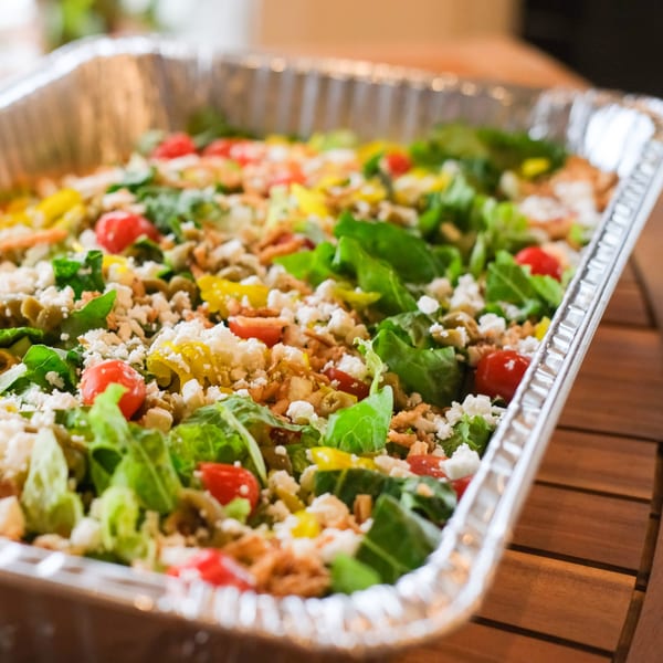 A large catering tray of fresh green salad with tomatoes, feta, and crispy toppings.
