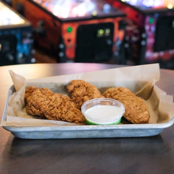 Crispy chicken tenders with ranch dipping sauce in a metal tray, with pinball machines in the background.