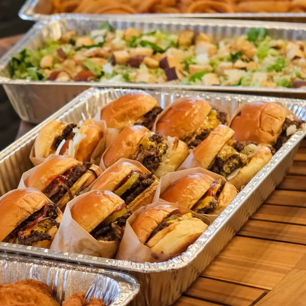 A catering tray of The Outsider's mini burgers with cheese and pickles, alongside a salad.