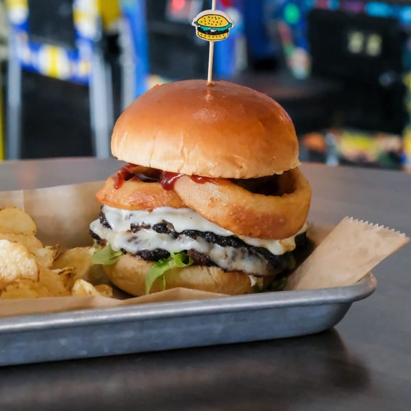 A double patty burger with onion rings, cheese, and lettuce, served with chips in a metal tray.