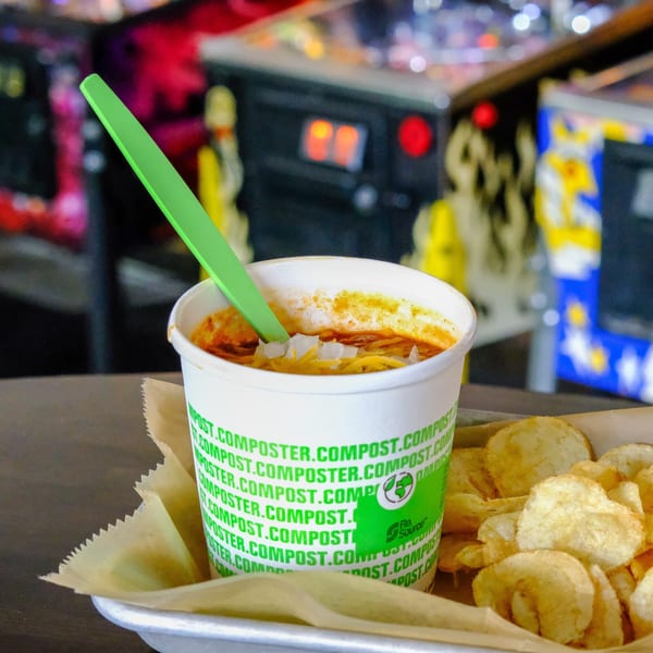 Chili with shredded cheese and diced onions in a compostable cup, served with potato chips.