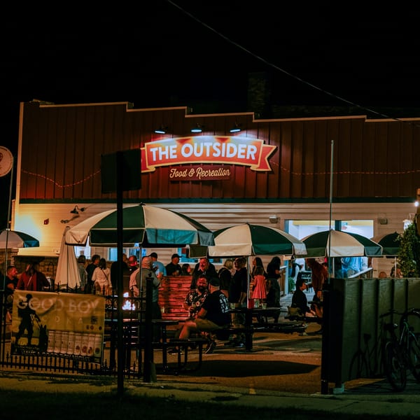 The Outsider restaurant at night, with a glowing sign and patrons gathered under umbrellas on the patio.