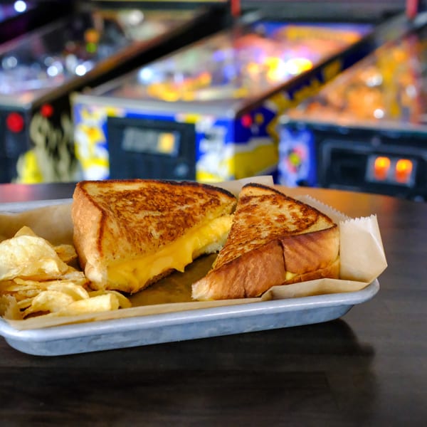 Grilled cheese sandwich with melted cheese and potato chips on a metal tray, with pinball machines in the background.