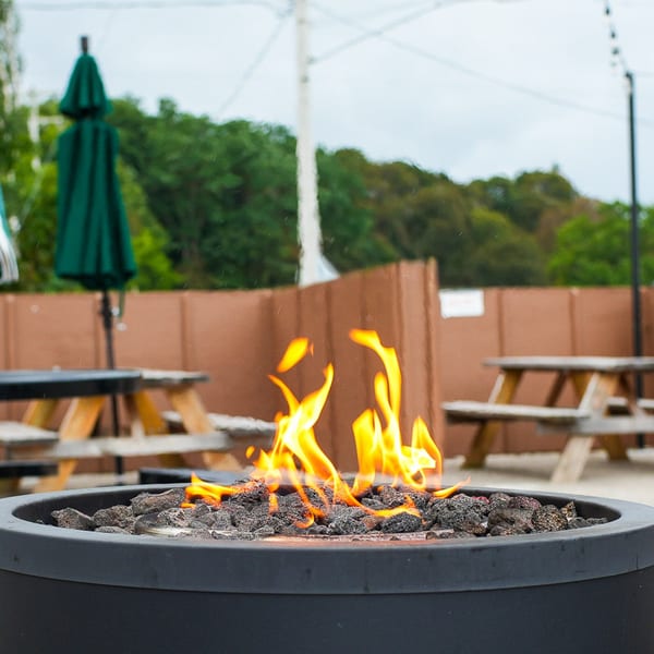Outdoor patio with a fire pit, picnic tables, and green umbrellas under a cloudy sky.