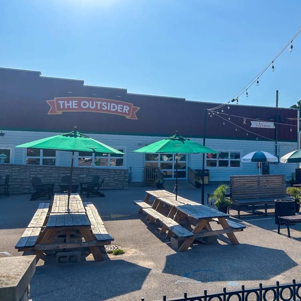 The Outsider restaurant exterior with a large patio featuring picnic tables, green umbrellas, and string lights.